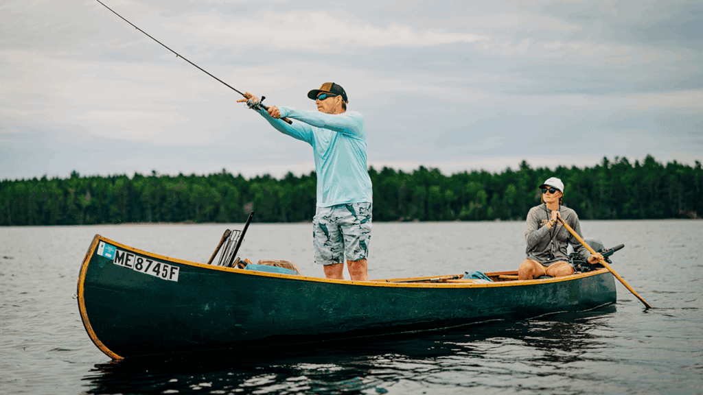 Two people in a green canoe on a lake; one stands and casts a fishing line while the other sits and paddles. Both wear casual outdoor clothing and hats, with a forest visible in the background.