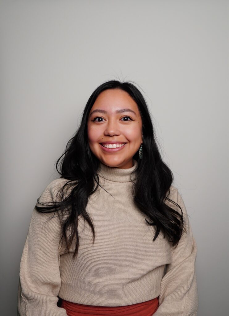 A woman with long dark hair, wearing a beige turtleneck sweater and orange skirt, smiles while sitting against a plain light gray background.