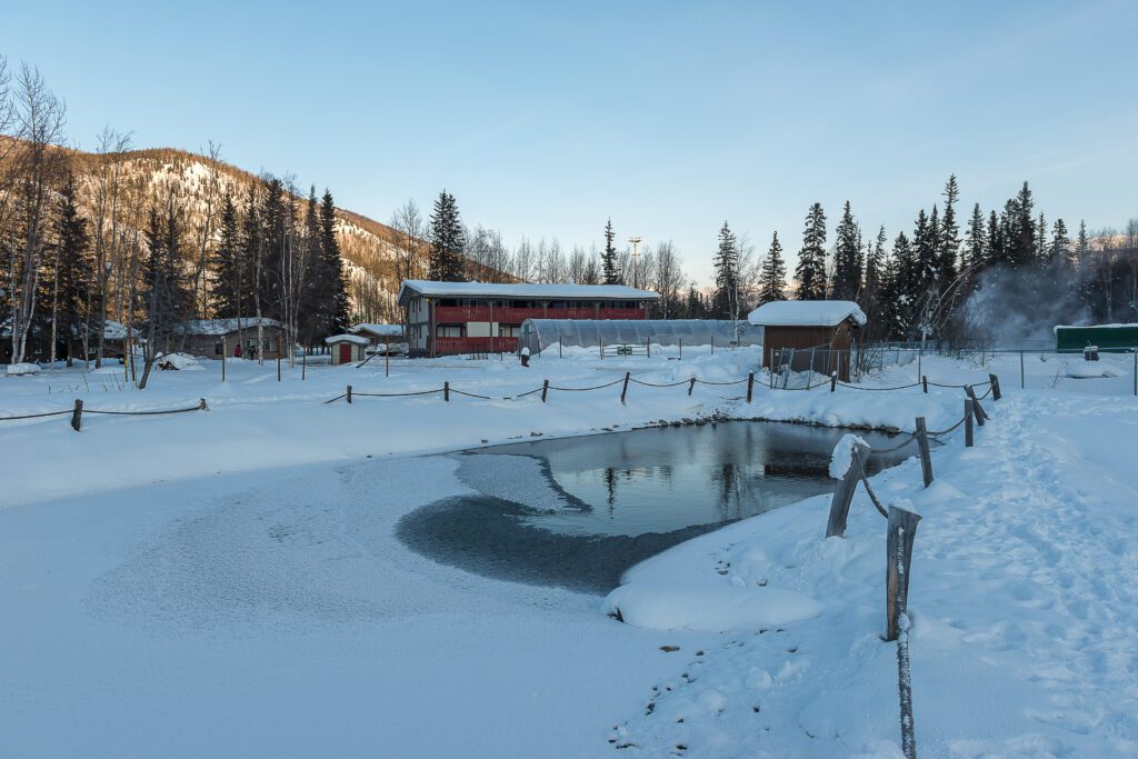 A partially frozen pond surrounded by snow with a red building, small cabins, and pine trees in the background on a clear winter day. A rope fence lines the snowy path beside the pond.