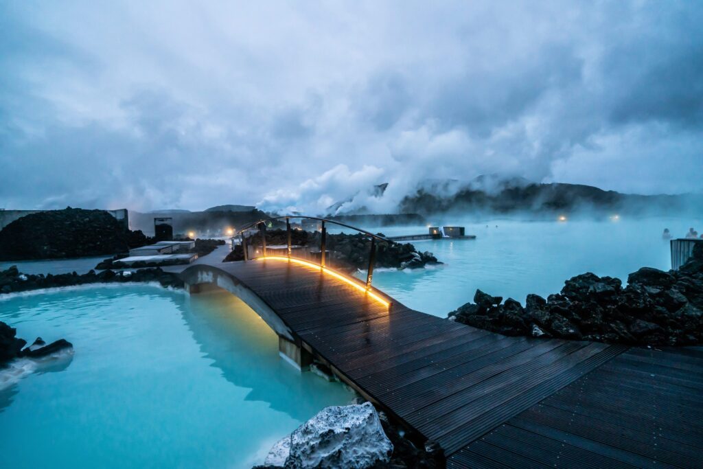 A wooden footbridge with glowing lights crosses over milky blue geothermal waters, surrounded by dark volcanic rocks and rising steam under a cloudy sky.