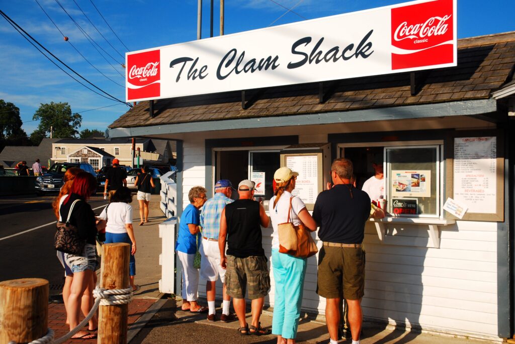 People stand in line outside The Clam Shack, a small white seafood stand with a menu board, under a bright sign with Coca-Cola logos, on a sunny day.