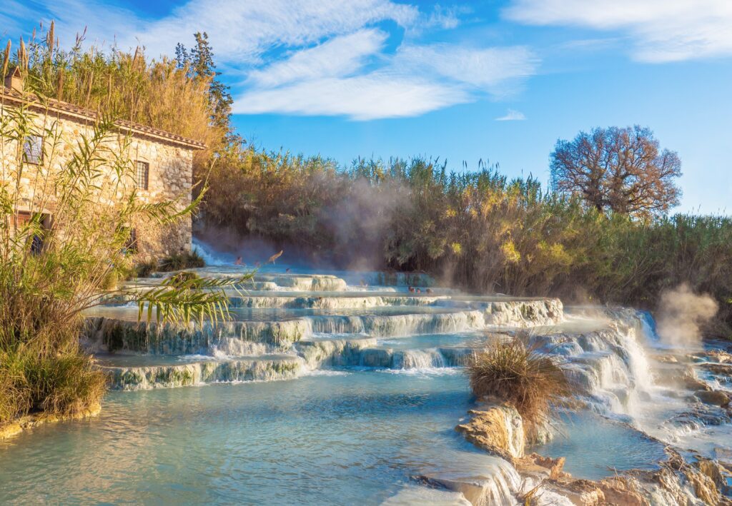 A terraced natural hot spring with steaming turquoise water flows beside a rustic stone building, surrounded by lush greenery and trees under a clear blue sky.