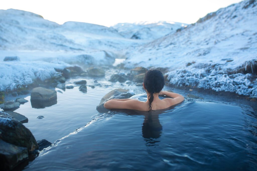 A person with dark hair is relaxing in a steaming natural hot spring surrounded by snowy mountains and rocks, with their back turned to the camera.