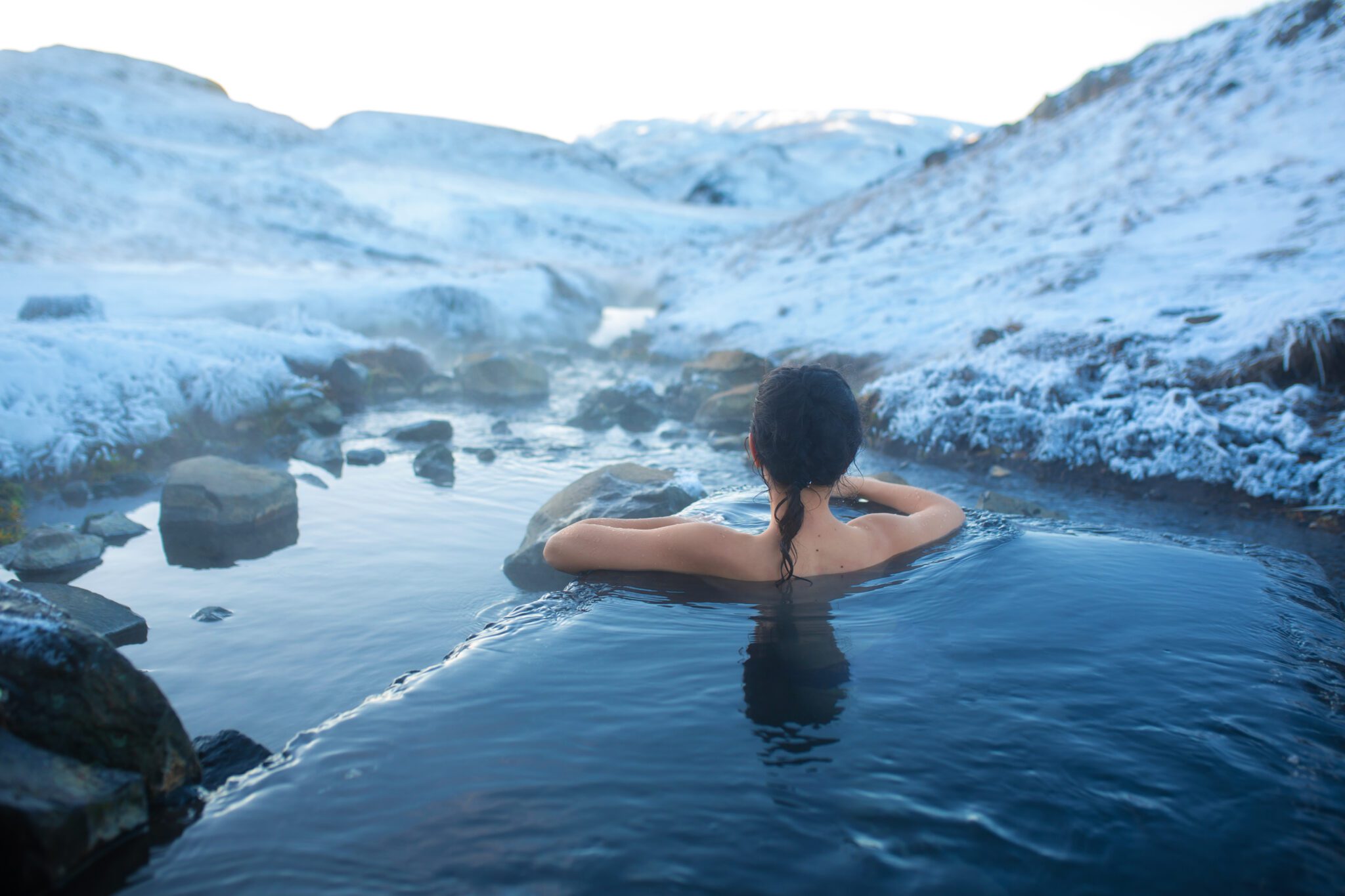 A person with dark hair is relaxing in a steaming natural hot spring surrounded by snowy mountains and rocks, with their back turned to the camera.