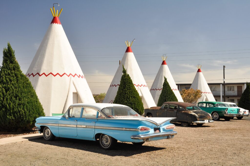 A row of white, teepee-shaped motel rooms with red zigzag trim, vintage cars parked in front, and green shrubs under a sunny sky.