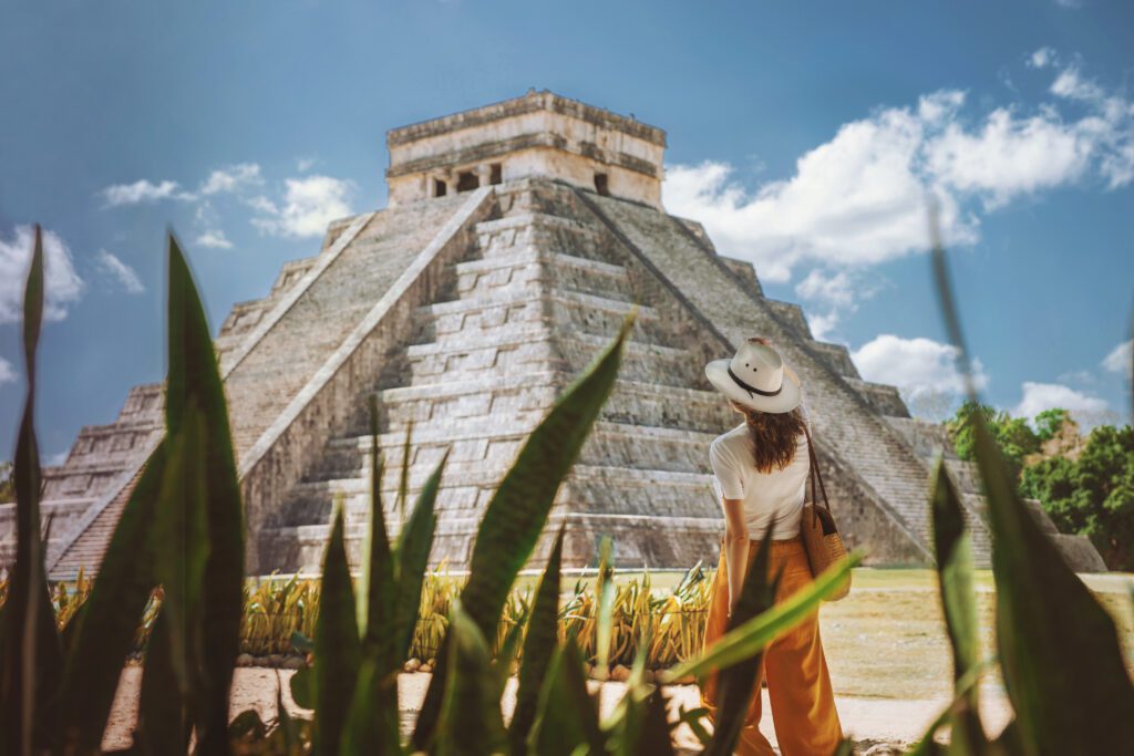A woman in a hat and orange pants stands facing the ancient pyramid of Chichen Itza in Mexico, with green plants in the foreground and a bright blue sky above.