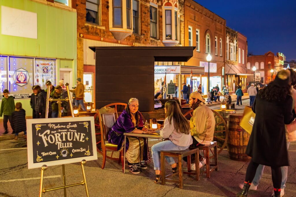 A fortune teller sits at a table with two people on a lively, brightly lit street at night. Nearby, a sign reads “Fortune Teller.” Pedestrians walk past storefronts in the background.