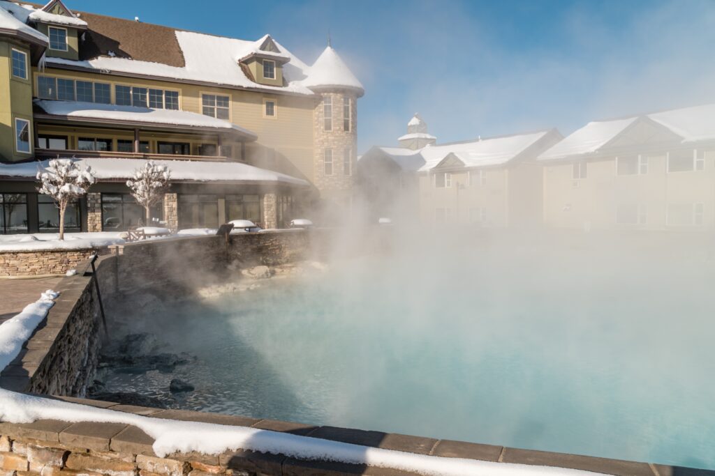 A steaming outdoor hot spring pool surrounded by snow and a large lodge-style building with snow-covered roofs under a clear blue sky.