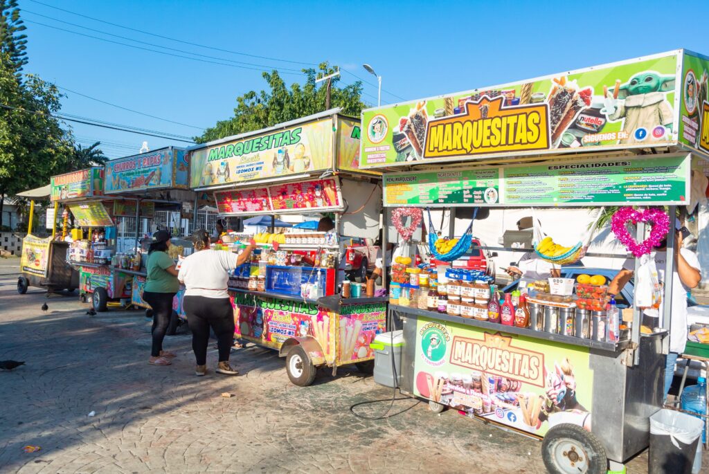 Colorful food carts selling snacks and drinks are lined up outdoors, with people ordering and preparing food. Bright signs and decorations cover the carts, and the scene is sunny with a clear blue sky overhead.