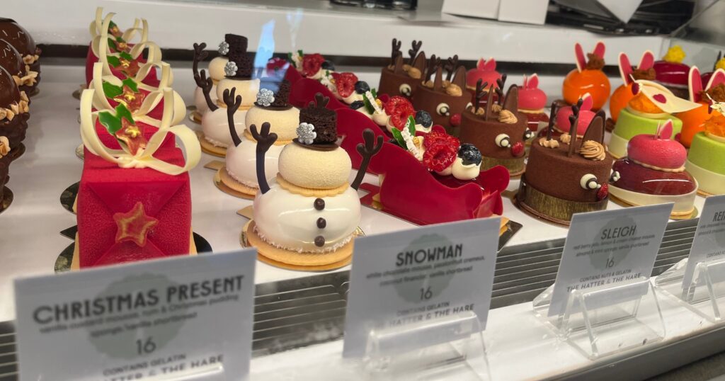A display case filled with festive holiday pastries, including white snowman-shaped cakes and chocolate cakes decorated as reindeer, along with other colorful desserts and labels in front of each treat.