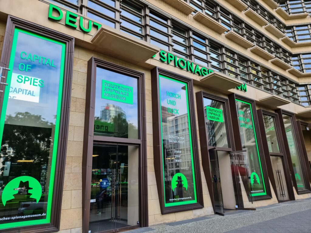 The exterior of the German Spy Museum in Berlin, with large green and blue signs in English and German on tall windows reading Capital of Spies and Deutsches Spionagemuseum.