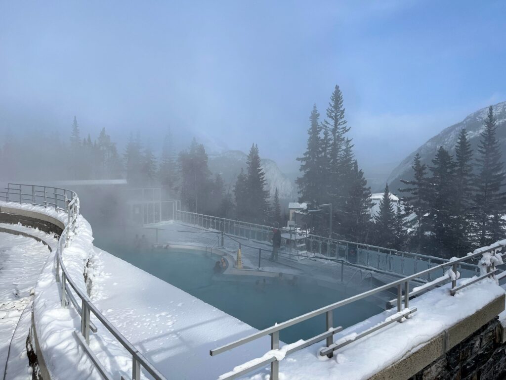 Steam rises from an outdoor hot spring pool surrounded by snow and pine trees, with mountains in the background. A few people are visible in the warm water, and railings are covered with snow. The sky is partly cloudy.