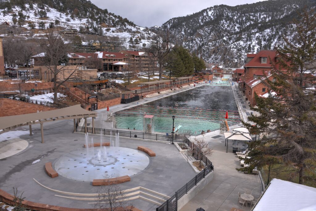 A large outdoor hot springs pool surrounded by snow-dusted buildings and mountains, with steam rising from the water and a circular fountain area in the foreground.