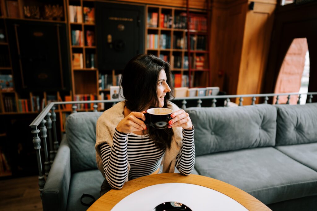 A woman with long dark hair, wearing a striped shirt and light sweater, sits on a gray couch in a cozy library café, holding a cup and looking to the side with a smile. Bookshelves fill the background.