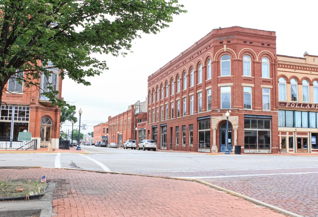 A quiet street corner in a small town with historic red brick buildings, large windows, and a tree in the foreground. Several parked cars line the street on a cloudy day.