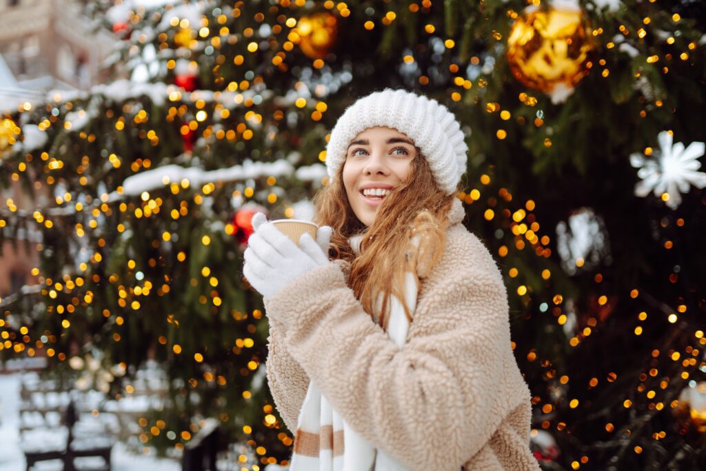 A young woman in winter clothes smiles and holds a cup, standing in front of a decorated Christmas tree with golden lights and ornaments outdoors.