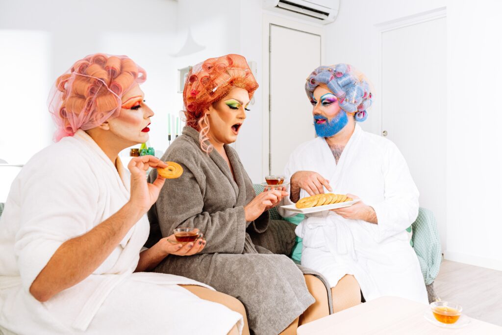 Three people wearing colorful wigs, makeup, and bathrobes sit together, eating cookies and drinking tea in a bright living room.