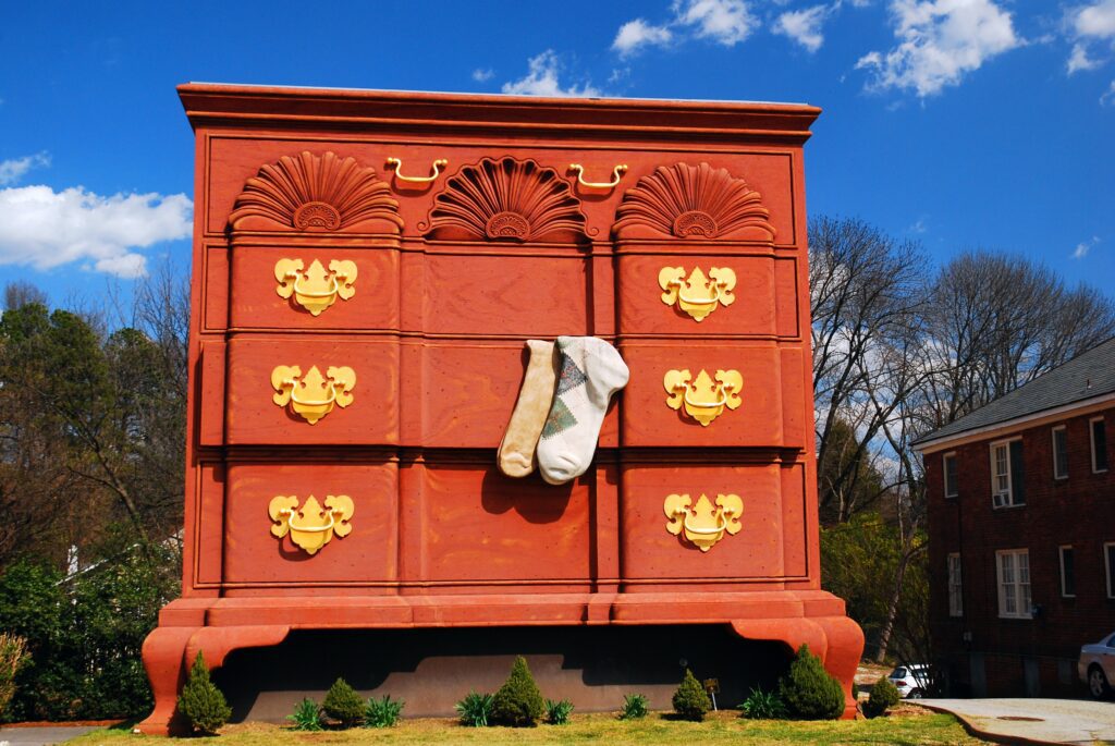 A giant red chest of drawers with oversized gold handles and two large socks hanging from a middle drawer stands outdoors near trees and a brick building.