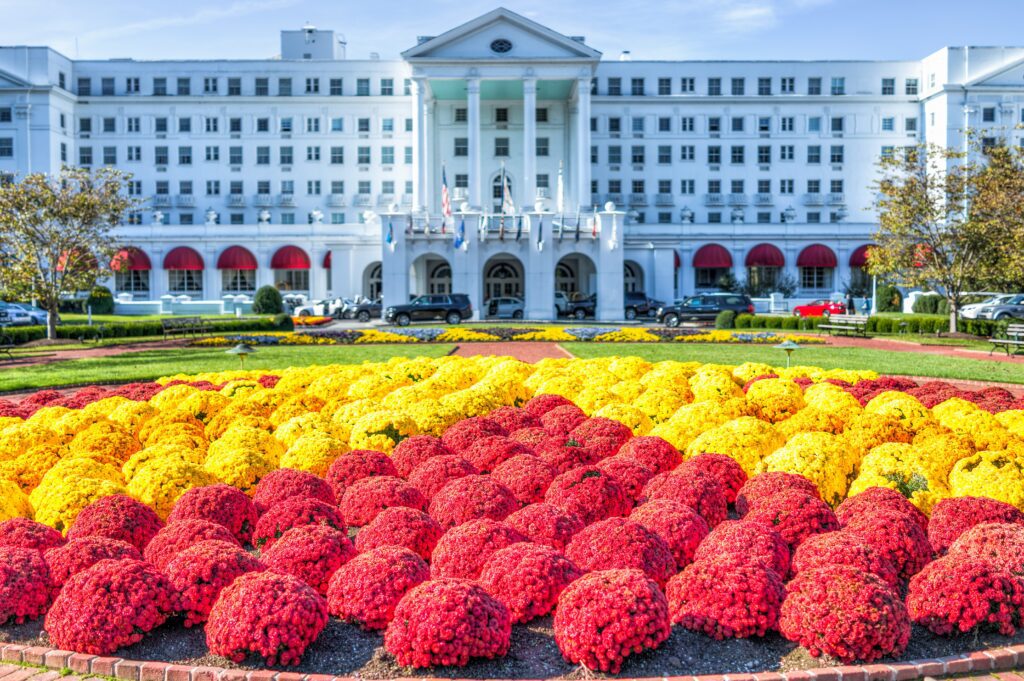 A large white hotel with red awnings is seen behind a colorful, well-maintained flower garden featuring red and yellow blooms.