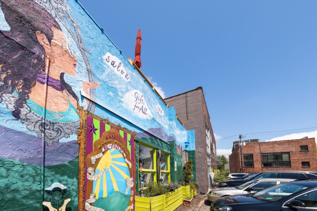 A colorful mural decorates the side of a building next to a parking lot with several cars under a clear blue sky.