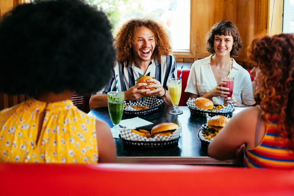 Four people sit at a diner table with burgers, fries, and drinks, smiling and talking with each other.