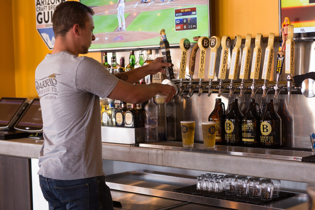 A bartender pours beer from a tap into a glass at a bar, with several draft beer handles and a TV showing a baseball game in the background.