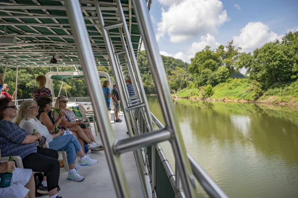 People sit on a covered boat as it travels along a calm river, with trees and a partly cloudy sky visible on the riverbank.