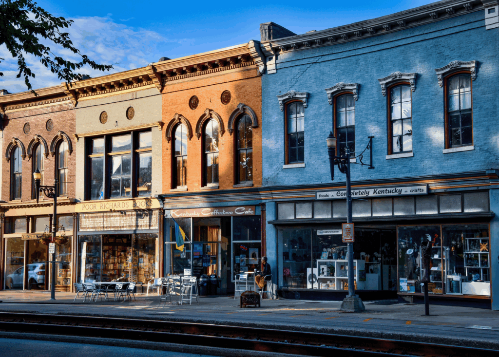 Three historic storefronts with large arched windows line a street. Sidewalk seating is outside a bookstore; other shops display crafts and gifts in their windows.