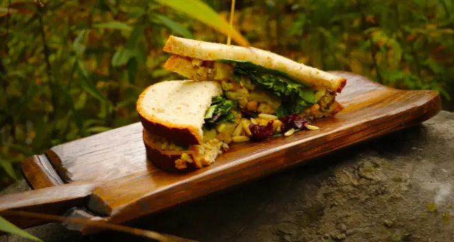 A sandwich with greens and what appears to be turkey and cranberries is cut in half and placed on a wooden board, outdoors with greenery in the background.