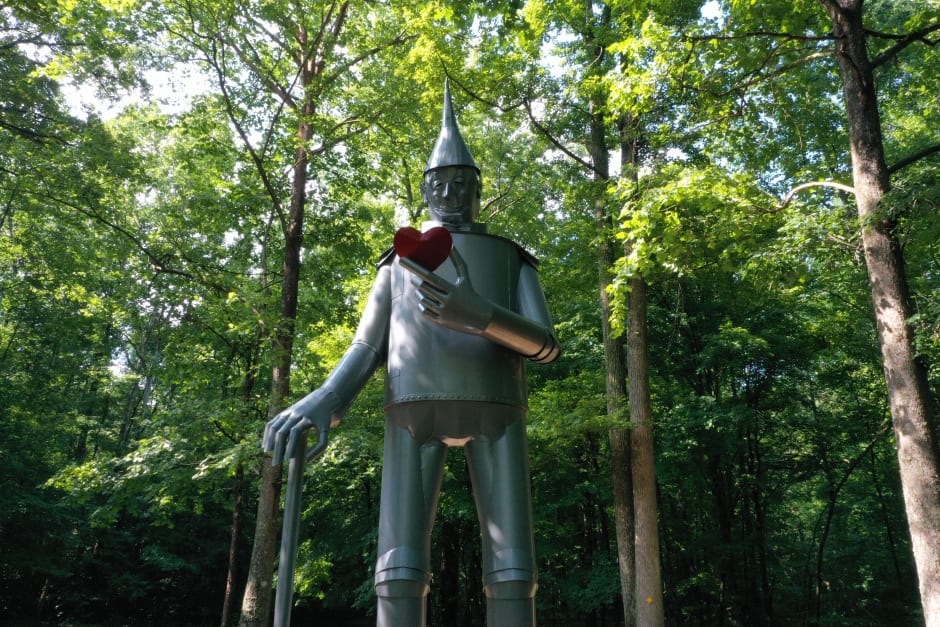 Large metal Tin Man statue holding a red heart stands among tall green trees in a sunlit wooded area.
