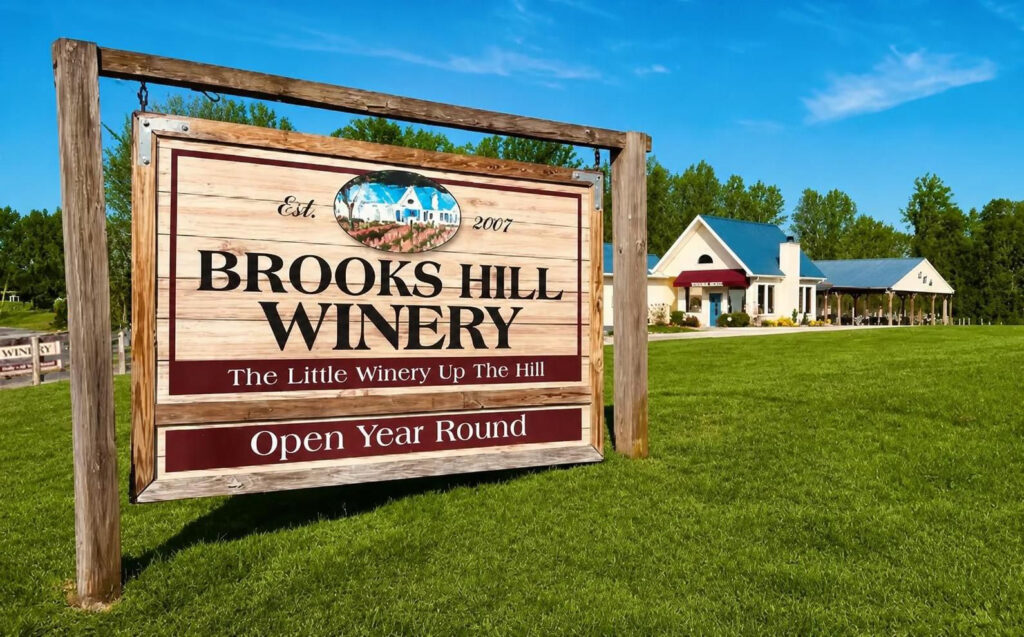 A wooden sign for Brooks Hill Winery stands on grass, with the winery building and trees visible in the background under a clear blue sky.