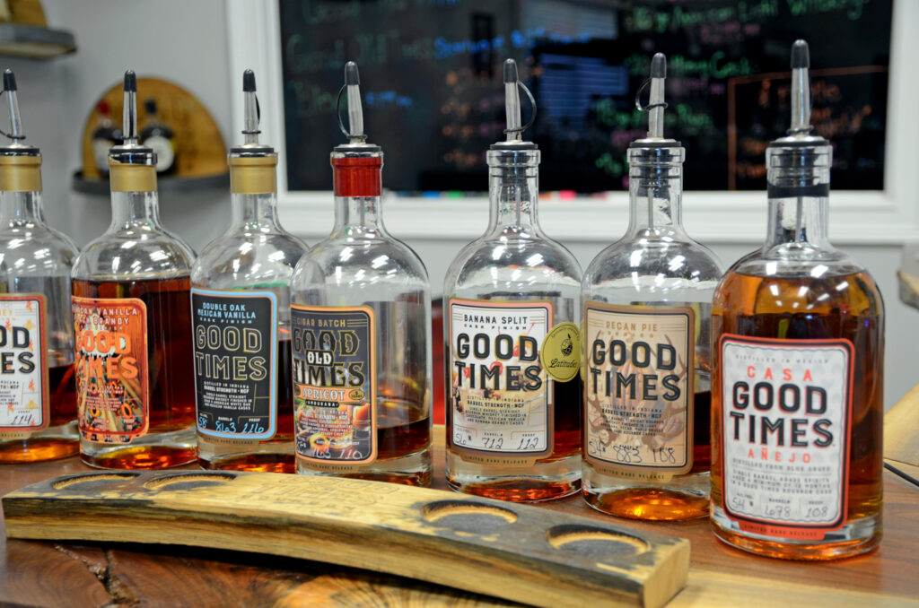 Seven bottles of Good Times whiskey with various labels are lined up on a wooden bar, with a tasting stave in front and a chalkboard in the background.