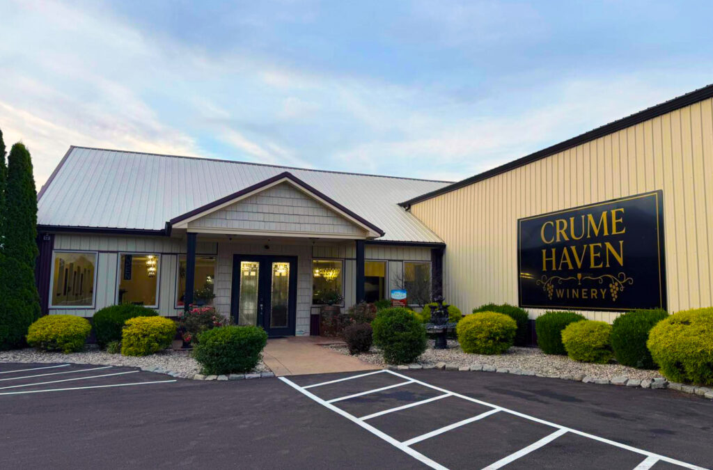 A beige building with a white roof and black sign reading “CRUME HAVEN WINERY” is surrounded by bushes, with a clear entrance and parking spaces in front.
