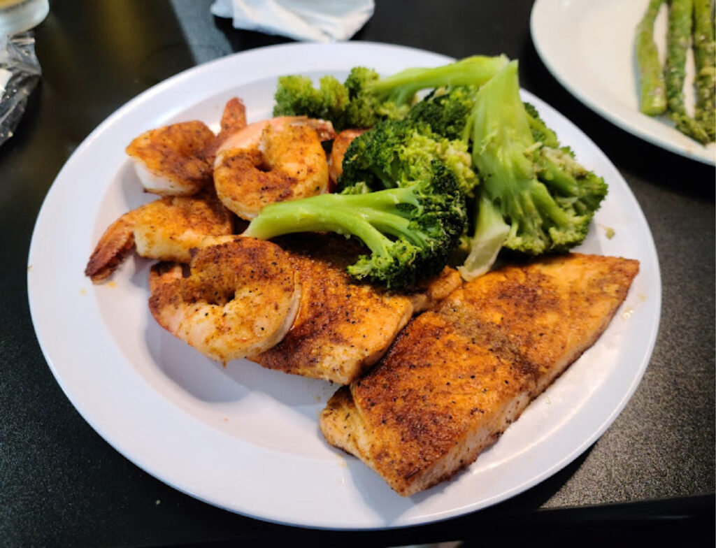 A white plate with seasoned grilled fish, shrimp, and steamed broccoli, served on a black table.