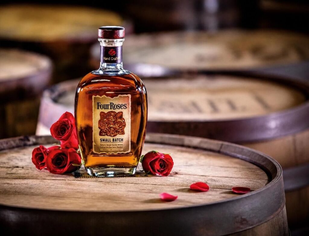 A bottle of Four Roses Small Batch bourbon sits on a wooden barrel, surrounded by three red roses and scattered rose petals, with more barrels visible in the blurred background.