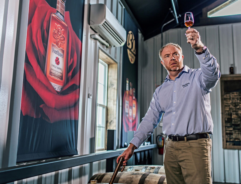 A man in a button-down shirt examines a glass of whiskey in a distillery, with a large rose and whiskey bottle poster on the wall behind him.
