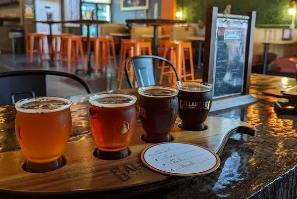 A wooden flight tray holds four small glasses of beer on a bar counter, with a tasting note card and bar seating visible in the background.