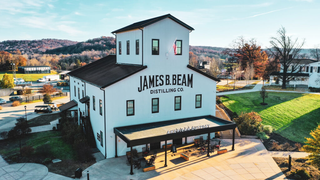 A white building labeled “James B. Beam Distilling Co.” sits on landscaped grounds under a clear sky, with “American Outpost” written above the entrance.