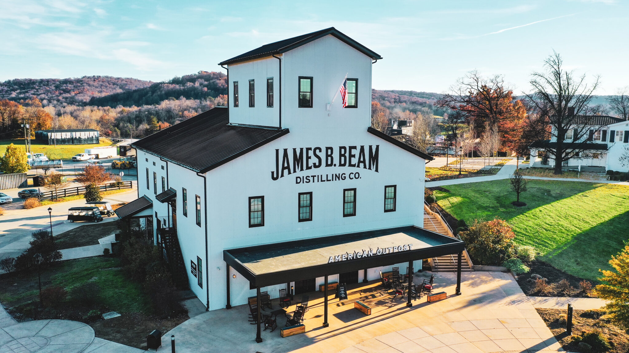 A white building labeled “James B. Beam Distilling Co.” sits on landscaped grounds under a clear sky, with “American Outpost” written above the entrance.
