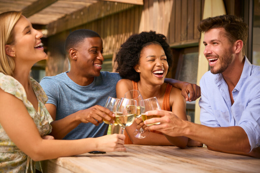 Four people sit at an outdoor table, smiling and clinking glasses of white wine together in a cheerful, social setting.