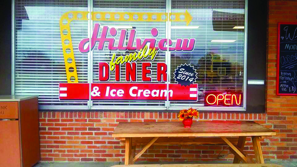 A storefront window displays a “Hillview Family Diner & Ice Cream” sign with “Open” lit up and a wooden picnic table with a small flower vase in front.