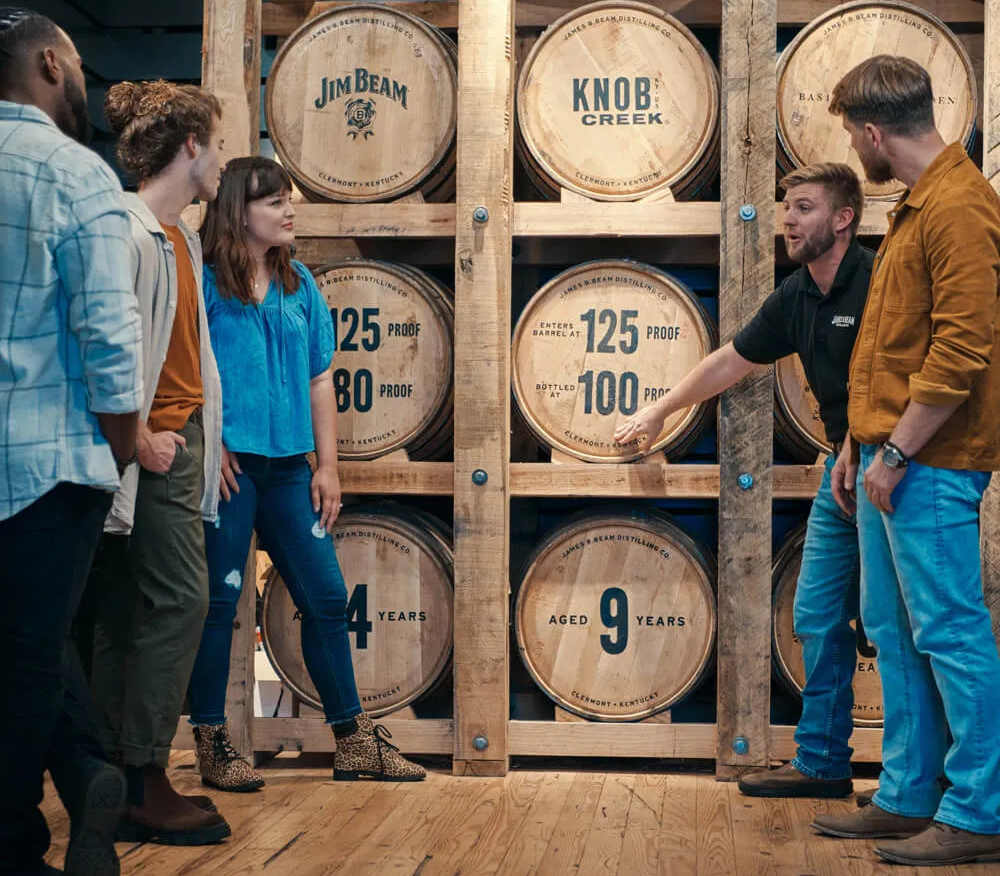 A group of four people listens to a guide in front of large stacked whiskey barrels labeled with brands, proof, and age in a distillery setting.