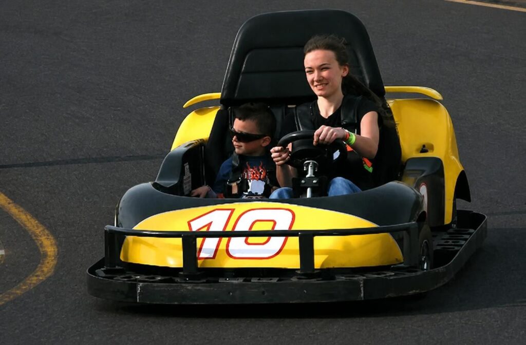 Two children ride in a yellow go-kart with the number 10 on the front, driving on a paved track.