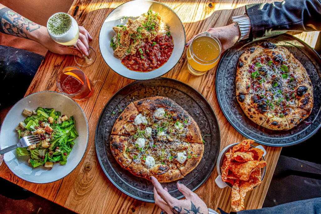 Two people share a meal at a wooden table with two pizzas, a pasta dish, a salad, chips, a beer, a cocktail, and a glass of a green-topped beverage.