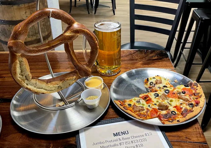 A large pretzel on a stand with sauces, a glass of beer, and a plate of pizza slices are on a wooden table next to a menu.
