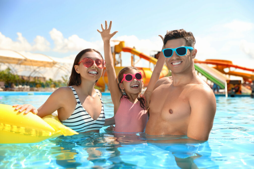A woman, a man, and a child wearing sunglasses are smiling and standing in a swimming pool with water slides in the background.