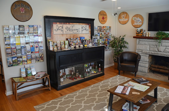 Visitor center interior with brochures on display, a welcome sign, liquor bottles in a cabinet, a fireplace, armchair, and tables with magazines on a patterned rug.