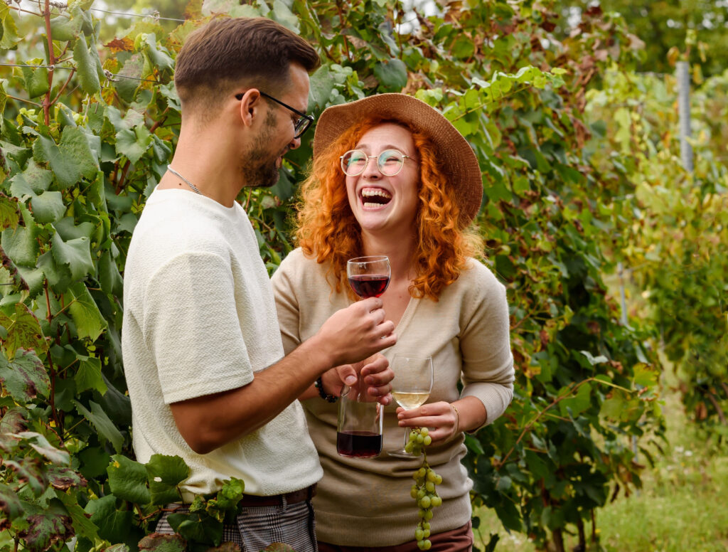 Two people stand among grapevines, smiling and holding wine glasses. One holds red wine, the other white, and a bunch of grapes.
