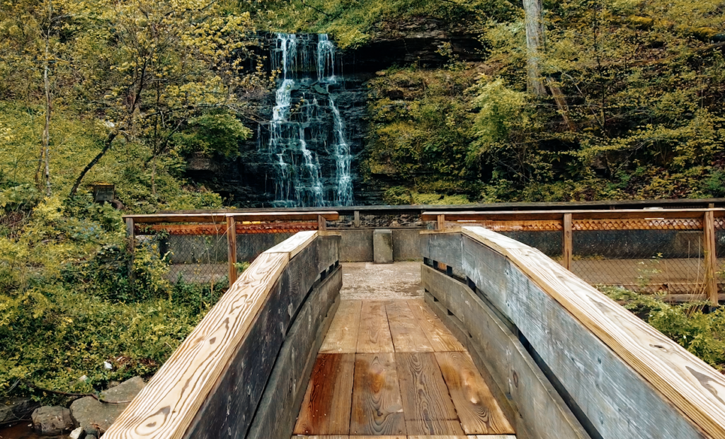 Wooden footbridge leading to a lookout platform with a fenced railing, overlooking a tall waterfall surrounded by green trees and foliage.
