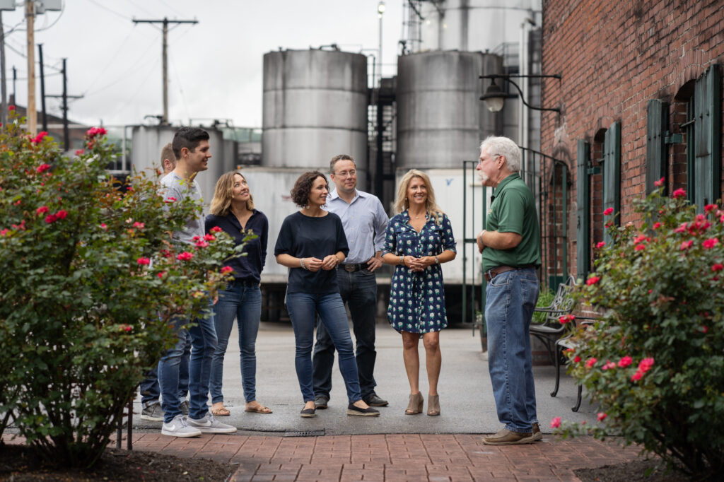 A group of six adults stand talking outdoors near brick buildings and industrial storage tanks, surrounded by flowering bushes.